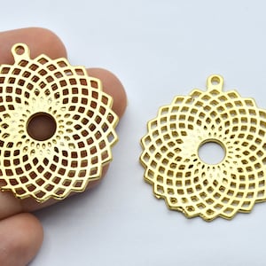 Pode incluir: Dois pingentes vazados dourados. Cada pingente tem formato circular com um furo central e um padrão geométrico repetitivo. Os pingentes têm uma pequena argola na parte superior para pendurar.