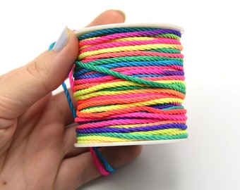 May include: A vibrant spool of multi-coloured nylon cord. The cord is tightly wound on a white plastic spool. The colours include pink, orange, yellow, green, blue, and purple. The cord is held by a hand against a white backdrop.