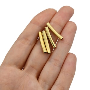 Puede incluir: Varios tubos metálicos pequeños de color dorado se sostienen en una mano sobre un fondo blanco. Los tubos son cilíndricos y parecen ser componentes para la fabricación de joyas u otras manualidades. Los tubos están cortados uniformemente y tienen un acabado brillante.