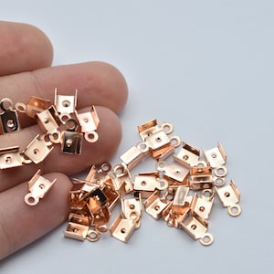 May include: A collection of small, copper-colored crimp end findings for jewelry making. Each finding has a rectangular body with a loop and two tabs for securing to a cord or ribbon. The findings are scattered on a light background.