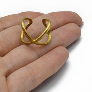 May include: A gold-colored ring with an overlapping X design. The ring is held in a hand against a white background. The ring appears to be made of metal and has a textured surface.
