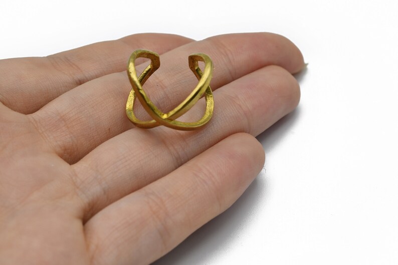 May include: A gold-colored ring with an overlapping X design. The ring is held in a hand against a white background. The ring appears to be made of metal and has a textured surface.
