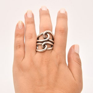 May include: Silver-toned ring with a unique, intertwined design. The ring features multiple loops and bands, creating a bold, textured look. The ring is worn on a hand with neutral-toned nail polish.