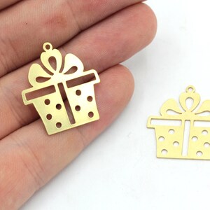 May include: Two gold-colored metal gift box charms. Each charm features a bow on top and dotted details on the box. The charms have a small loop at the top for attaching to jewelry or other crafts.
