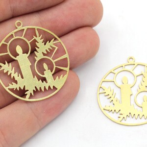 May include: Two gold-colored metal pendants. Each pendant is a circle with a cutout design featuring two candles, fir branches, and a sunburst pattern. The pendants have a small loop at the top for hanging.