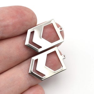 May include: Two silver-colored geometric charms with a hexagonal shape and a cutout design. Each charm has a small loop at the top for attaching to jewelry. The charms are displayed on a white background.