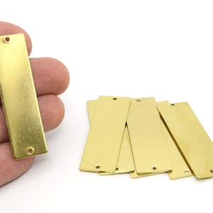 Puede incluir: Una colección de etiquetas rectangulares de metal de latón con esquinas redondeadas y dos pequeños agujeros. Una etiqueta es sostenida por una mano, mientras que otras están dispersas sobre una superficie blanca. Las etiquetas tienen un acabado dorado brillante.