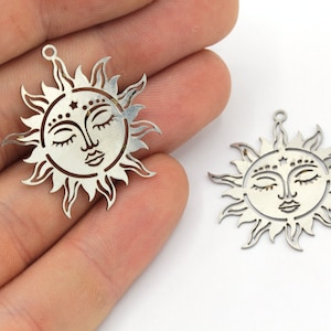 Peut inclure: Deux breloques solaires argent&eacute;es avec des yeux ferm&eacute;s et des traits d&eacute;taill&eacute;s. Chaque breloque a une boucle en haut pour suspendre et un motif d&eacute;coratif de rayons de soleil autour du visage. Sur fond blanc.