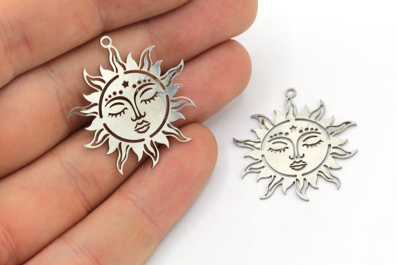 Peut inclure: Deux breloques solaires argent&eacute;es avec des yeux ferm&eacute;s et des traits d&eacute;taill&eacute;s. Chaque breloque a une boucle en haut pour suspendre et un motif d&eacute;coratif de rayons de soleil autour du visage. Sur fond blanc.