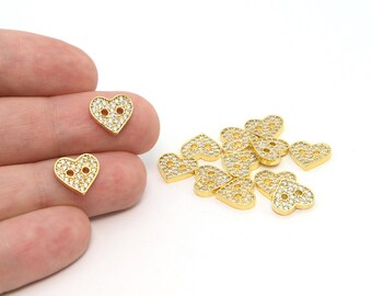 May include: Gold-colored heart-shaped buttons with clear crystal accents. The buttons have two holes for sewing and are scattered on a white surface. The buttons are approximately 0.5 inches in size.