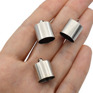 May include: Three silver-colored cylindrical metal components with a small loop on top, resting in a person's open palm. The cylinders have a smooth, reflective surface and a dark interior.