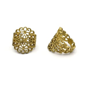 May include: Two gold-colored filigree rings are displayed against a white background. The rings feature an openwork design with floral and geometric patterns. One ring is upright, while the other is tilted.