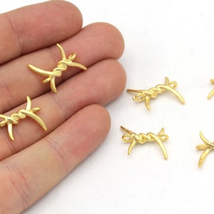 May include: Gold-colored metal barbed wire charms. The charms are small, with a twisted center and curved barbs extending outwards. The charms are displayed against a white background.