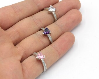 May include: Three silver rings with heart-shaped gemstones in white, purple, and pink. The ring bands are adorned with small, clear stones. The rings are displayed on a hand against a white background.