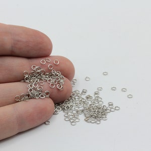 May include: A collection of small, silver-colored jump rings, scattered on a white surface and held in a hand. These open rings are used for jewelry making and other crafts. The rings are approximately 0.25 inches in diameter.