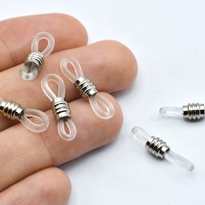 May include: Clear eyeglass retainers with silver metal accents. The retainers have a loop design for attaching to glasses and a cylindrical metal piece. Several are shown on a white background.