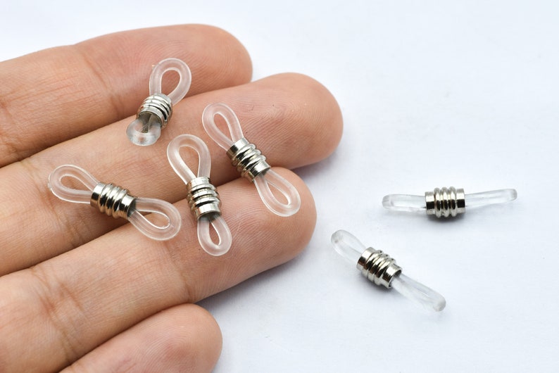 May include: Clear eyeglass retainers with silver metal accents. The retainers have a loop design for attaching to glasses and a cylindrical metal piece. Several are shown on a white background.
