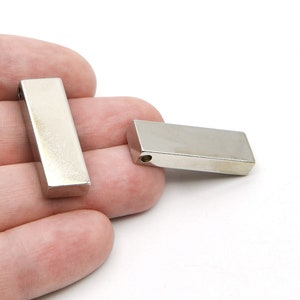 Könnte beinhalten: Zwei rechteckige, silberfarbene Metallanhänger mit poliertem Finish. Jeder Anhänger hat ein kleines Loch, das durch ein Ende gebohrt wurde. Die Anhänger werden vor einem weißen Hintergrund gezeigt.