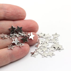 May include: A collection of small, silver-colored star charms with a hole at the top for stringing. The charms are scattered on a white surface, with some held in a hand. These charms are ideal for jewelry making or craft projects.