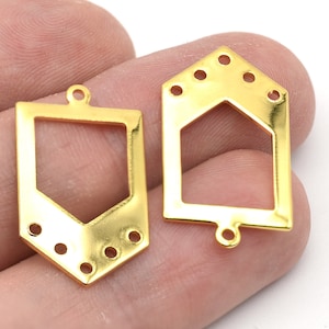 Może przedstawiać: Dwa złote metalowe elementy biżuteryjne. Jeden ma kształt tarczy z prostokątnym otworem i spiczastym dnem. Drugi ma kształt domu z sześciokątnym otworem. Oba mają otwory do mocowania.