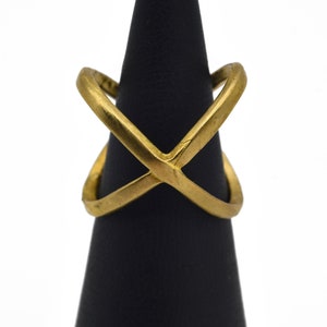 May include: A gold-colored ring with an X-shaped design. The ring is displayed on a black stand against a white background. The ring's design features two intersecting bands, creating an X shape. The ring is made of a shiny, metallic material.