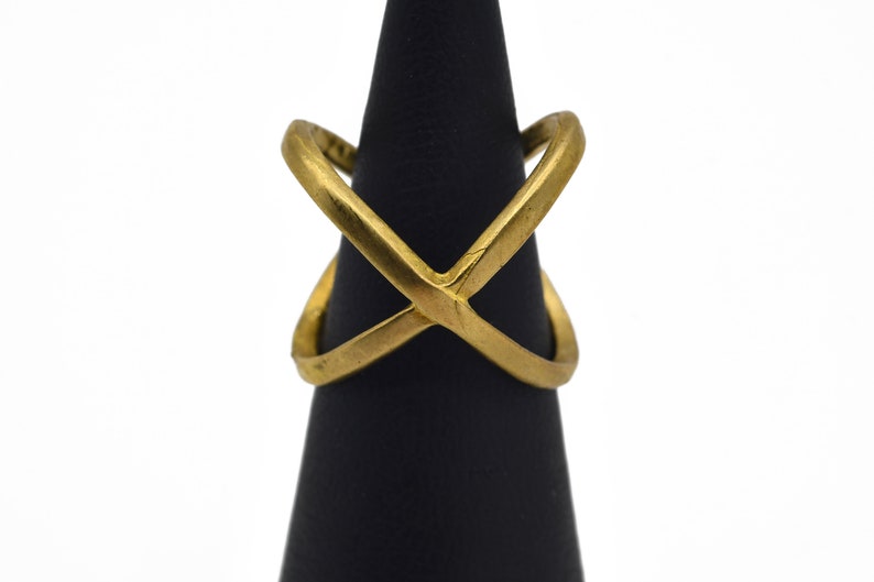 May include: A gold-colored ring with an X-shaped design. The ring is displayed on a black stand against a white background. The ring's design features two intersecting bands, creating an X shape. The ring is made of a shiny, metallic material.