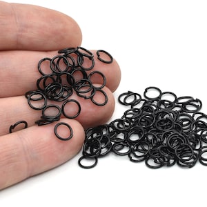 May include: A collection of small, black metal jump rings, scattered on a white surface. The rings are open and circular, suitable for jewelry making or crafts. The image is a close-up, showing the details of the rings.