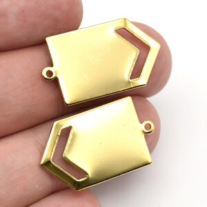 May include: Two gold-colored metal pendants. Each pendant has a rectangular shape with a cut-out arrow design on one side and a small loop at the top for attaching to a chain or other jewelry component. The pendants are shown against a white background.