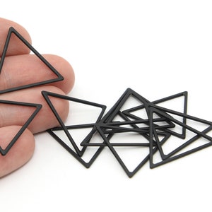 May include: A selection of black metal triangle charms. The charms are open-framed and have a matte finish. Some are scattered, while others are held in a hand. The background is white.