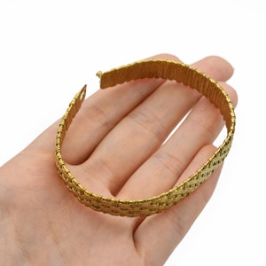 May include: A gold-colored bracelet with a woven design. The bracelet is held open, showing the clasp. The woven pattern gives the bracelet a textured appearance. The bracelet is approximately 0.75 inches wide.
