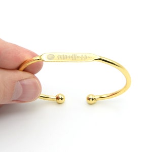 May include: A gold-colored cuff bracelet with a rectangular bar featuring a music waveform design. The bracelet has a smooth, polished finish and is held by a hand against a white background. The ends of the bracelet have small, round, gold-colored accents.