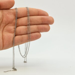 May include: A delicate silver-colored chain necklace with a fine, linked design. The necklace is displayed against a plain white background, showcasing its simple elegance. The chain is held in a hand, highlighting its length and detail.