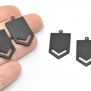 Könnte beinhalten: Nahaufnahme mehrerer schwarzer Metallanhänger. Jeder Anhänger hat eine rechteckige Form mit einem ausgeschnittenen Chevron-Design am unteren Rand und einer kleinen Schlaufe oben zum Befestigen an einer Kette oder einem anderen Schmuckbestandteil.