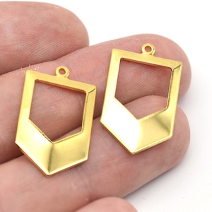 Może przedstawiać: Dwa złote geometryczne wisiorki o prostokątnym kształcie i spiczastym dnie. Każdy wisiorek ma małą pętlę u góry do przymocowania do łańcuszka lub innego elementu biżuterii. Wisiorki są trzymane w dłoni.