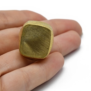 Puede incluir: Un anillo cuadrado de color dorado con un acabado de metal cepillado. El anillo se sostiene en una mano sobre un fondo blanco. El diseño del anillo es simple y moderno, con una parte superior ligeramente redondeada.