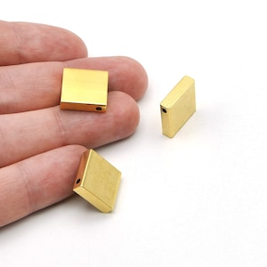 Puede incluir: Tres pequeñas cuentas cuadradas de metal dorado con un agujero perforado en cada una. Las cuentas se muestran sobre un fondo blanco. Las cuentas son probablemente para la fabricación de joyas u otros proyectos de artesanía.