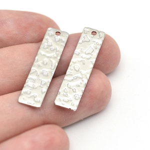Pode incluir: Dois pingentes retangulares de metal prateado com superfície texturizada e um pequeno orifício na parte superior. Os pingentes são segurados em uma mão contra um fundo branco. Estes pingentes são adequados para fazer joias.