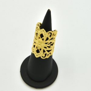 May include: A gold-colored filigree ring with an openwork floral design. The ring is displayed on a black ring holder against a white background. The ring's intricate details are highlighted by the light.