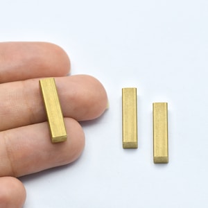 May include: Close-up of three small, rectangular, brass-colored metal bars. One bar is held in a hand, while the other two are on a white surface. The bars have a matte finish and are evenly cut.