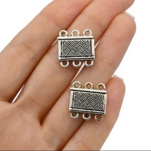 Puede incluir: Dos cierres de joyería rectangulares plateados con un patrón tejido. Cada cierre tiene tres bucles en cada extremo para sujetar a las joyas. Los cierres se sostienen en la mano de una persona.