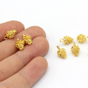 Può includere: Ciondoli in metallo color oro a forma di piccole pigne. I ciondoli hanno una superficie strutturata e un anello per il fissaggio ai gioielli. Diversi ciondoli sono sparsi su una superficie bianca.