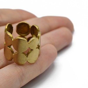 Op de afbeelding: Een goudkleurige ring met een herhalend patroon van verbonden cirkels. De ring is breed en heeft een getextureerde, matte afwerking. Het ontwerp heeft open ruimtes tussen de cirkels, waardoor een uniek, opengewerkt effect ontstaat. De ring wordt getoond op een hand.