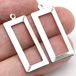 May include: Two silver-colored rectangular pendant blanks with a loop at the top for jewelry making. The pendants have a hollow center and a polished finish, perfect for crafting earrings or necklaces.