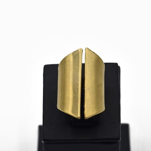 Op de afbeelding: Een goudkleurige ring met een gesplitst ontwerp. De ring wordt gepresenteerd op een zwarte standaard tegen een witte achtergrond. Het ontwerp van de ring heeft twee verticale, gebogen, rechthoekige vormen die aan de basis zijn verbonden.