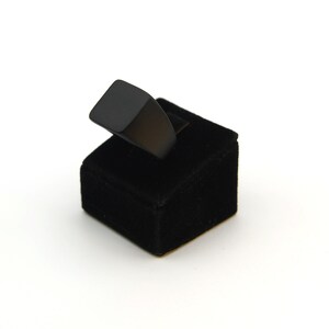 May include: A black ring with a square face sits on a small black velvet ring display. The ring has a minimalist design. The background is plain white.