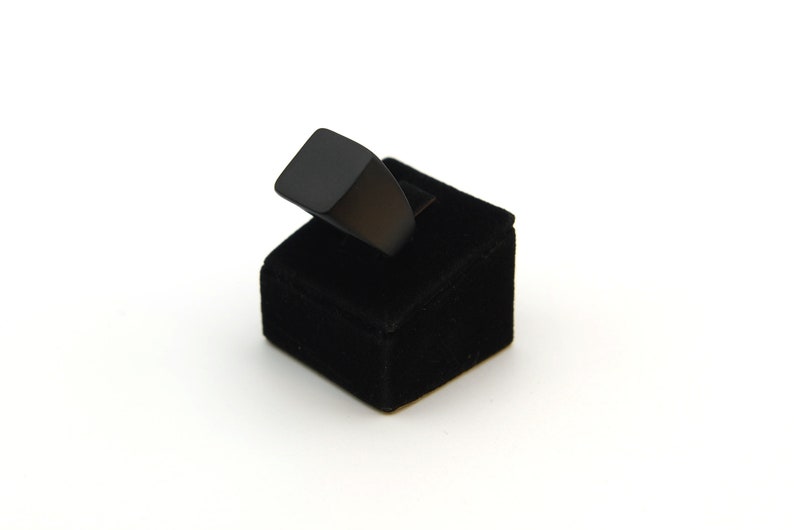 May include: A black ring with a square face sits on a small black velvet ring display. The ring has a minimalist design. The background is plain white.
