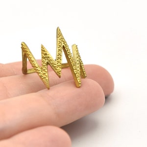 以下が含まれることがあります： 幾何学模様のゴールドカラーのリング。リングは、大胆でモダンな美しさを生み出す、一連の角度のあるジグザグの形が特徴です。白い背景を背景に、手に持たれています。