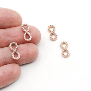 Könnte beinhalten: Anhänger mit Unendlichkeitszeichen aus Roségold mit klaren Kristallakzenten. Die Anhänger sind wie das Unendlichkeitszeichen geformt, mit einem doppelten Schlaufendesign. Die Kristalle sind um die Ränder gesetzt.
