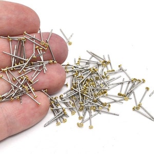 May include: A collection of small, silver-colored metal pins with gold-colored heads. The pins are scattered on a white surface. These pins are likely used for jewelry making or other craft projects.