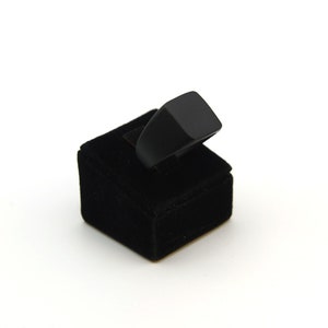 May include: A black ring with a square top is displayed on a black velvet ring holder. The ring has a simple, modern design. The ring holder is a small, square block.